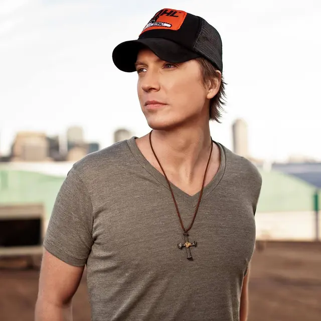 Brett Gillan in a trucker cap and grey T-shirt with a city skyline behind him